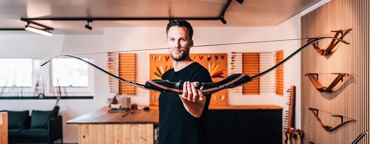 BEARPAW Store Rossach – Experte präsentiert einen hochwertigen Langbogen, umgeben von Bogenkollektion an der Wand, ideal für Beratung und Tests nahe Coburg.